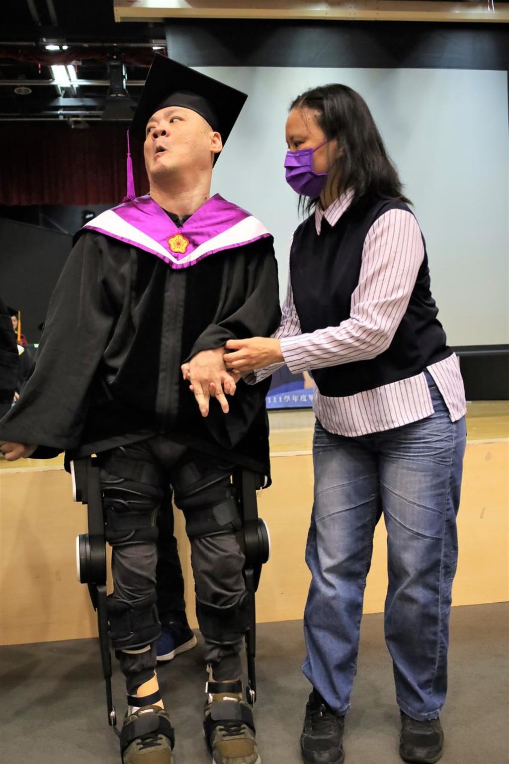 亞大腦麻博士生陳銘哲(左)，穿著「外骨骼機器人」，人生第1次從輪椅站起來，能夠行走，放開大姊陳秀玉手去領獎。圖／亞洲大學提供